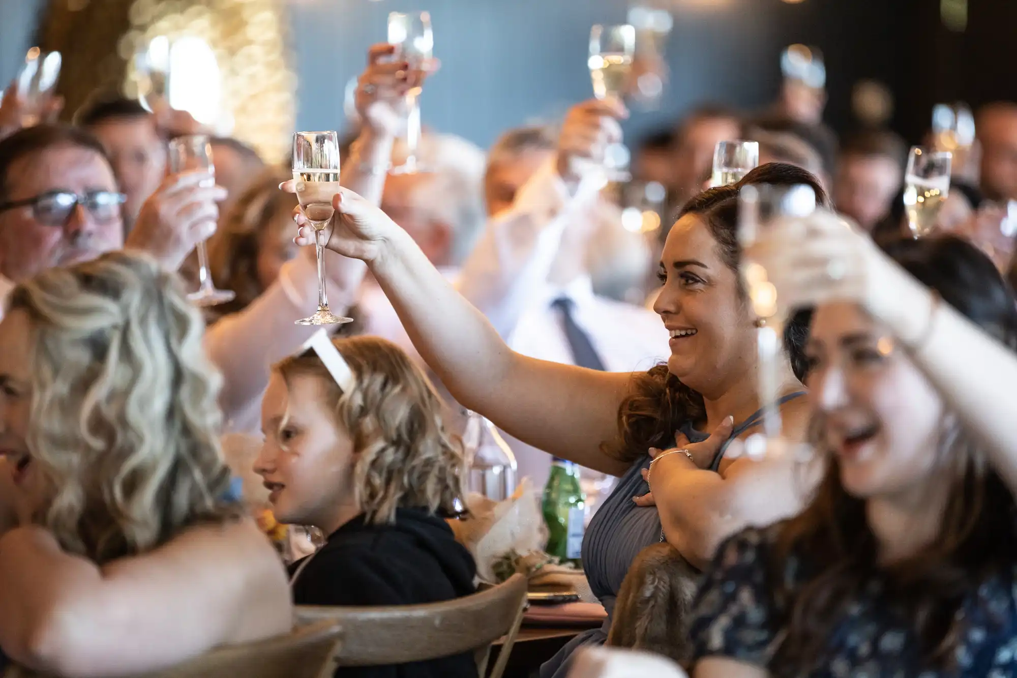 A group of people seated at tables raise their glasses in a toast, smiling and appearing to celebrate at an indoor event.