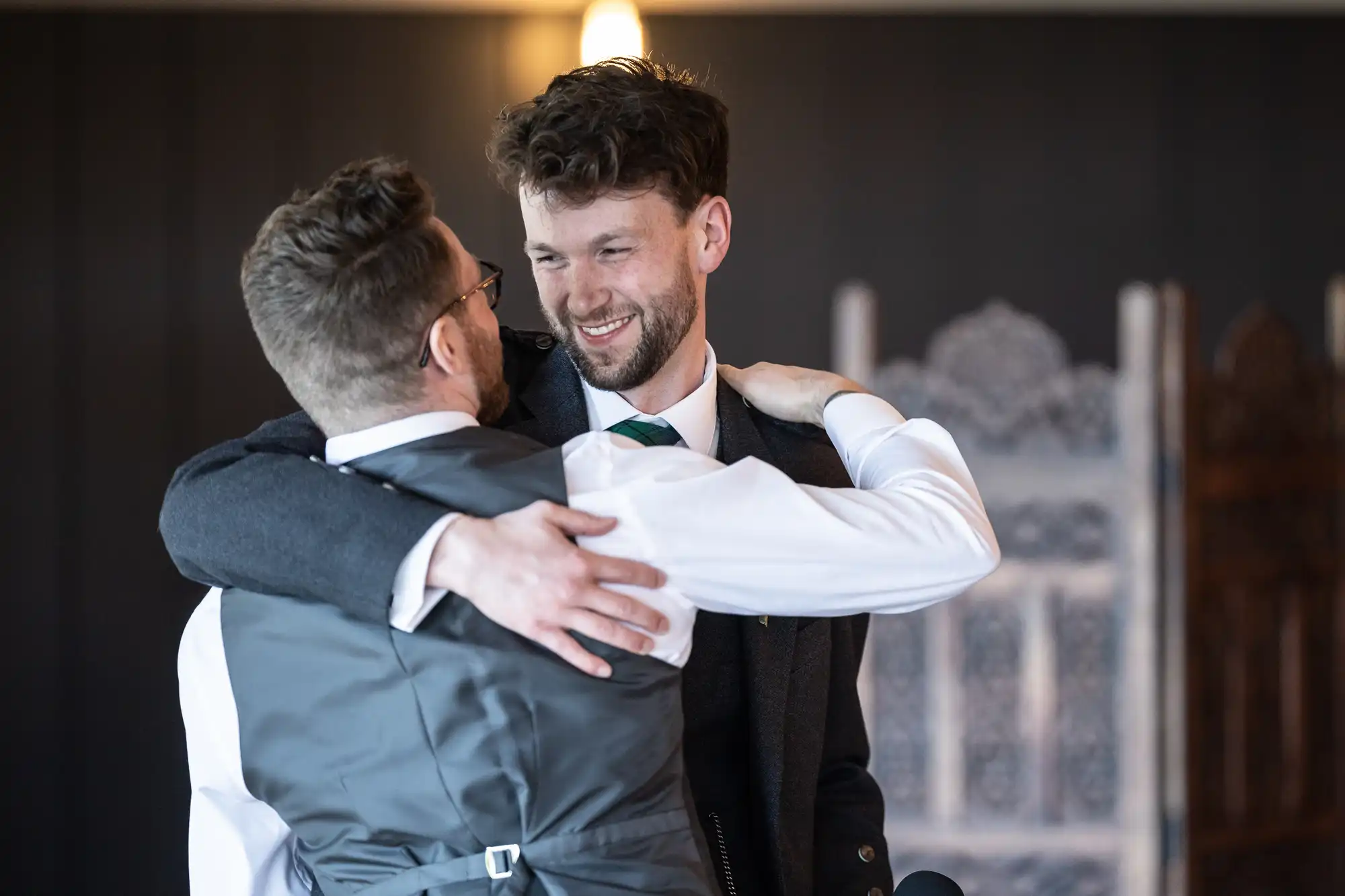 Two men embrace warmly indoors, smiling at one another.