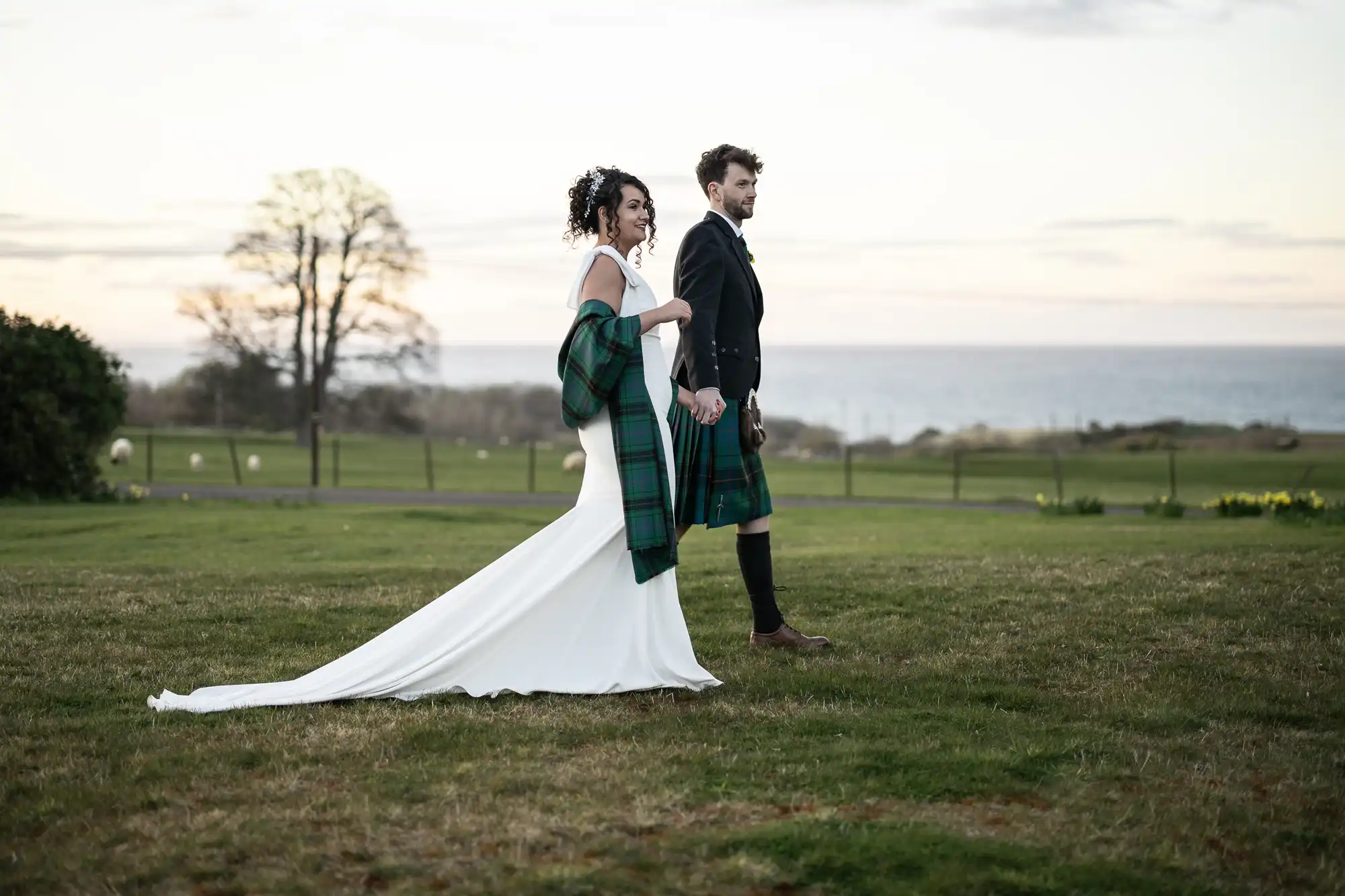 A couple dressed in wedding attire, the man in a kilt and the woman in a white dress, walk hand-in-hand on a grassy field near the seaside.