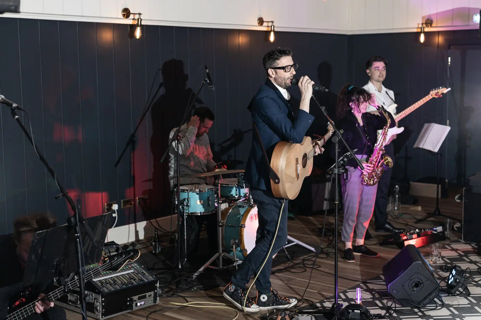 A band performs on stage with members playing guitar, drums, saxophone, and bass. The guitarist is also singing into a microphone. The stage is dimly lit with spotlights.