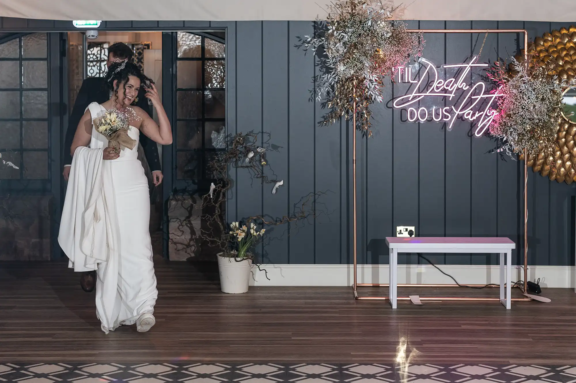 A bride in a white dress holding a bouquet walks alongside a groom in a dark suit. They enter a room with a neon sign stating "Til Death Do Us Party" and a decorative flower arrangement.