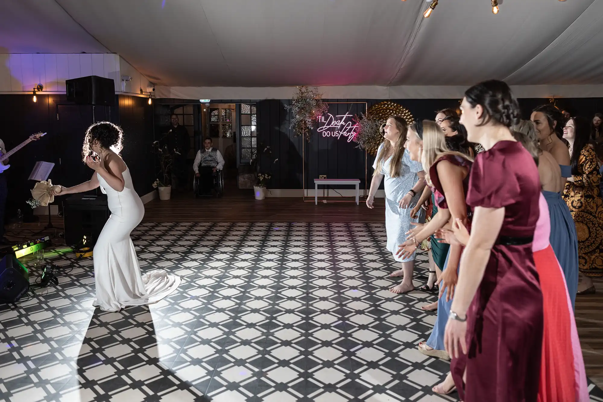 A bride throws her bouquet towards a group of women, who are eagerly waiting to catch it on a checkered floor inside a decorated hall.