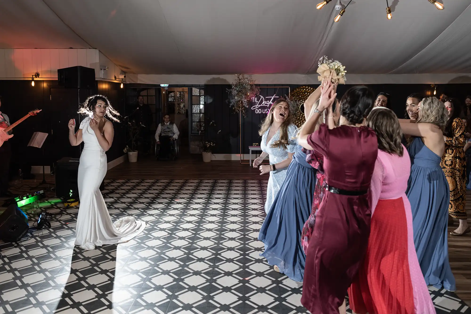 A bride in a white dress dances while a group of women await the bouquet toss in a room with patterned tile flooring and overhead lighting.