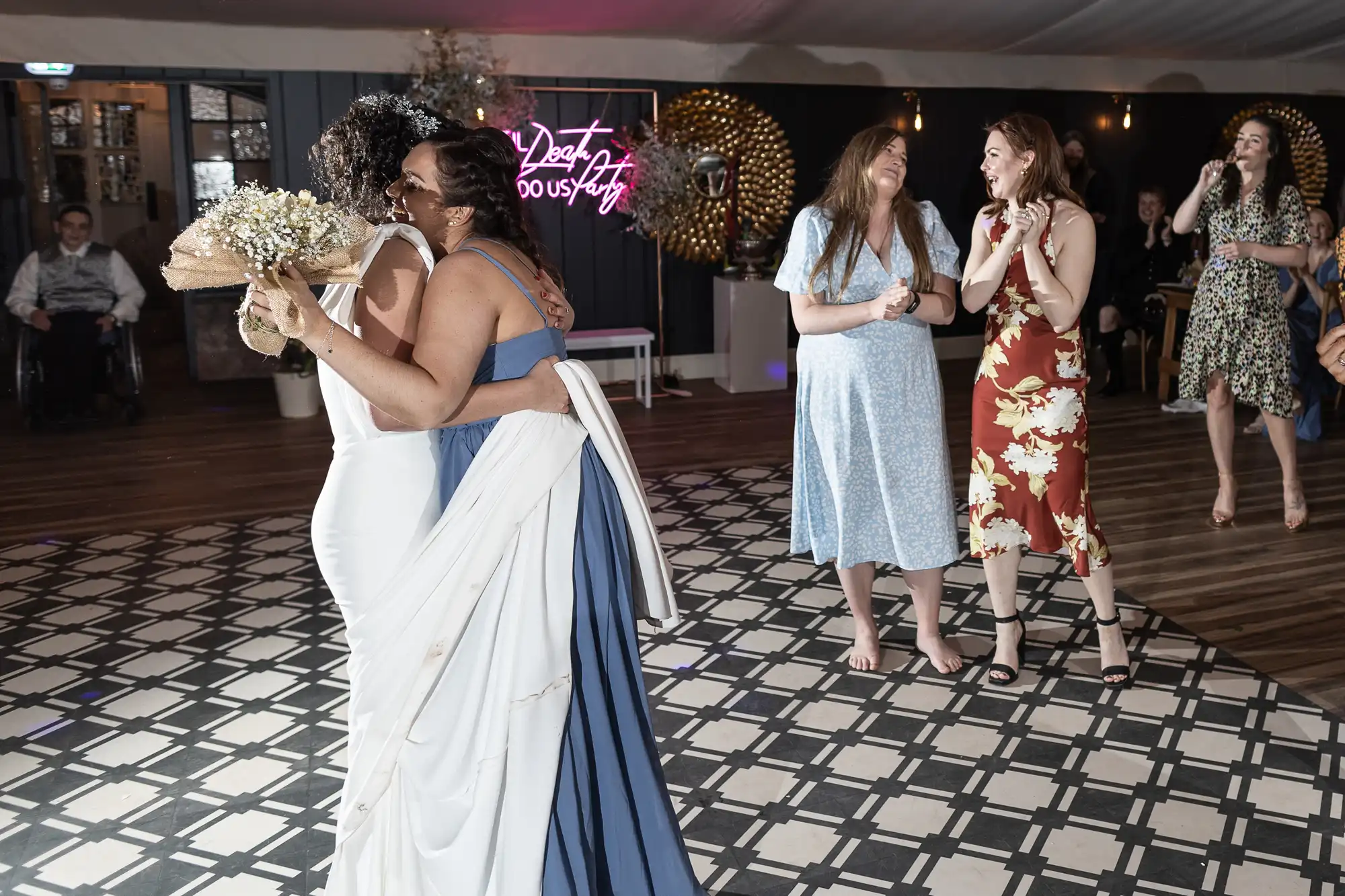 Two women embrace, one in a white dress holding a bouquet. Three other women look on, standing nearby on a checkered floor with a "Best Day Ever" sign in the background.