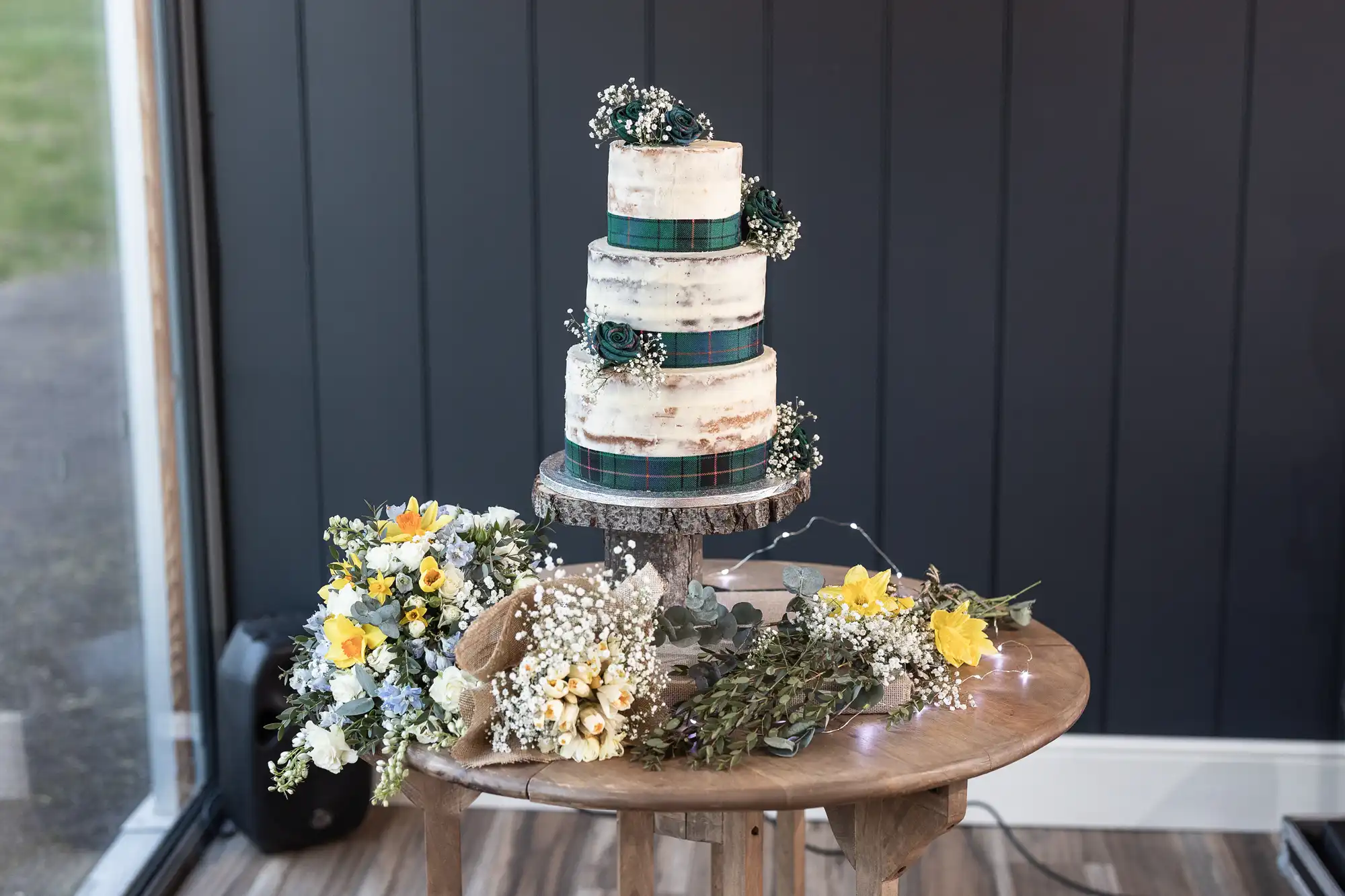 Three-tiered white cake with green plaid ribbons, adorned with small white flowers, placed on a wooden table alongside floral bouquets of yellow, white, and blue flowers. Dark wall background.