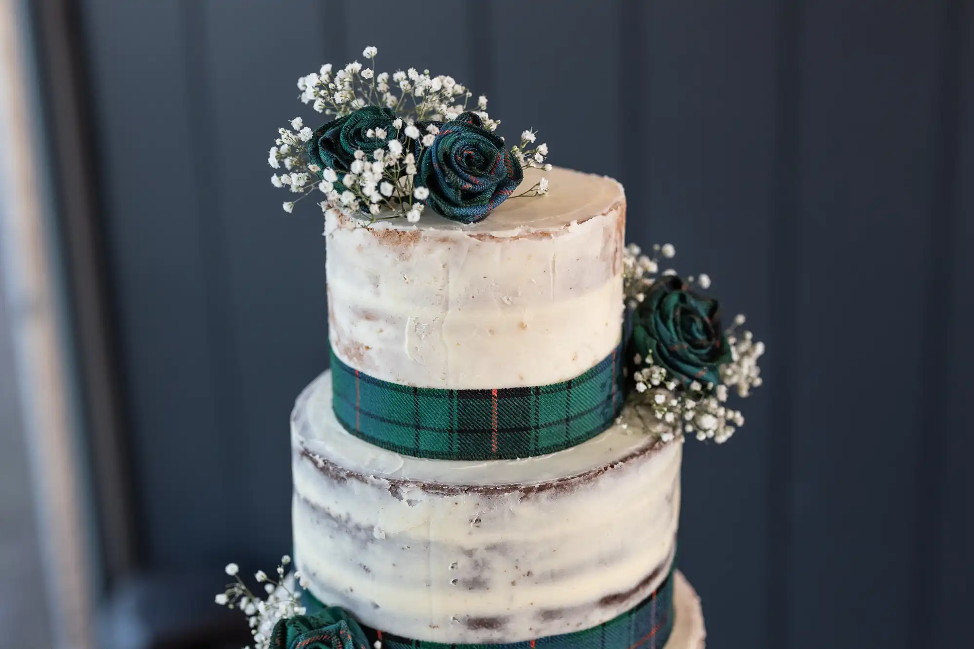 A three-tier semi-naked cake decorated with green tartan ribbon, artificial green roses, and sprigs of baby's breath.