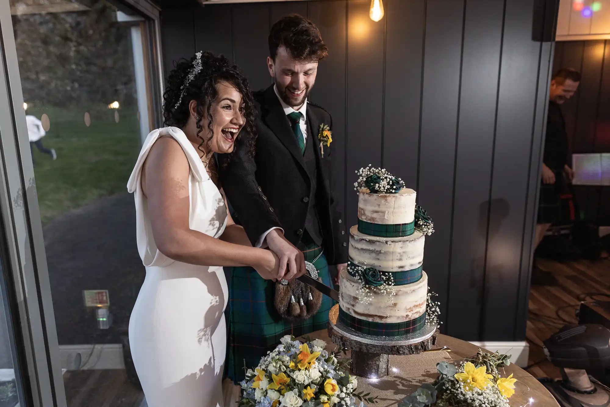 A couple in wedding attire smiles while cutting a three-tiered cake. The groom wears a kilt, and the bride is in a white dress. Two floral arrangements are on the table beside the cake.