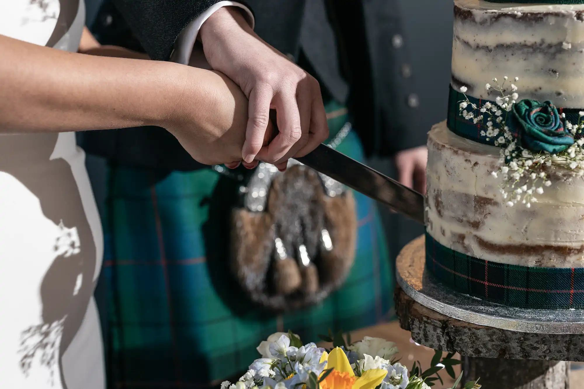 A couple, dressed in traditional Scottish attire, cuts a multi-tiered rustic cake decorated with flowers and greenery.