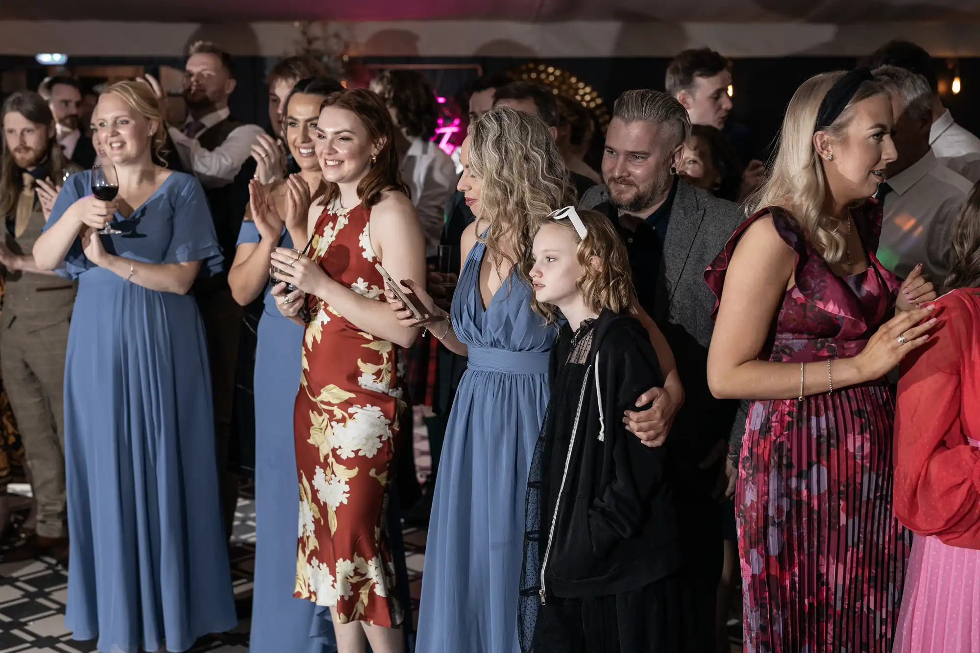 A group of people, some clapping and some standing together, watch an event. Several women wear dresses, and one child is present among the group. They appear attentive and engaged.