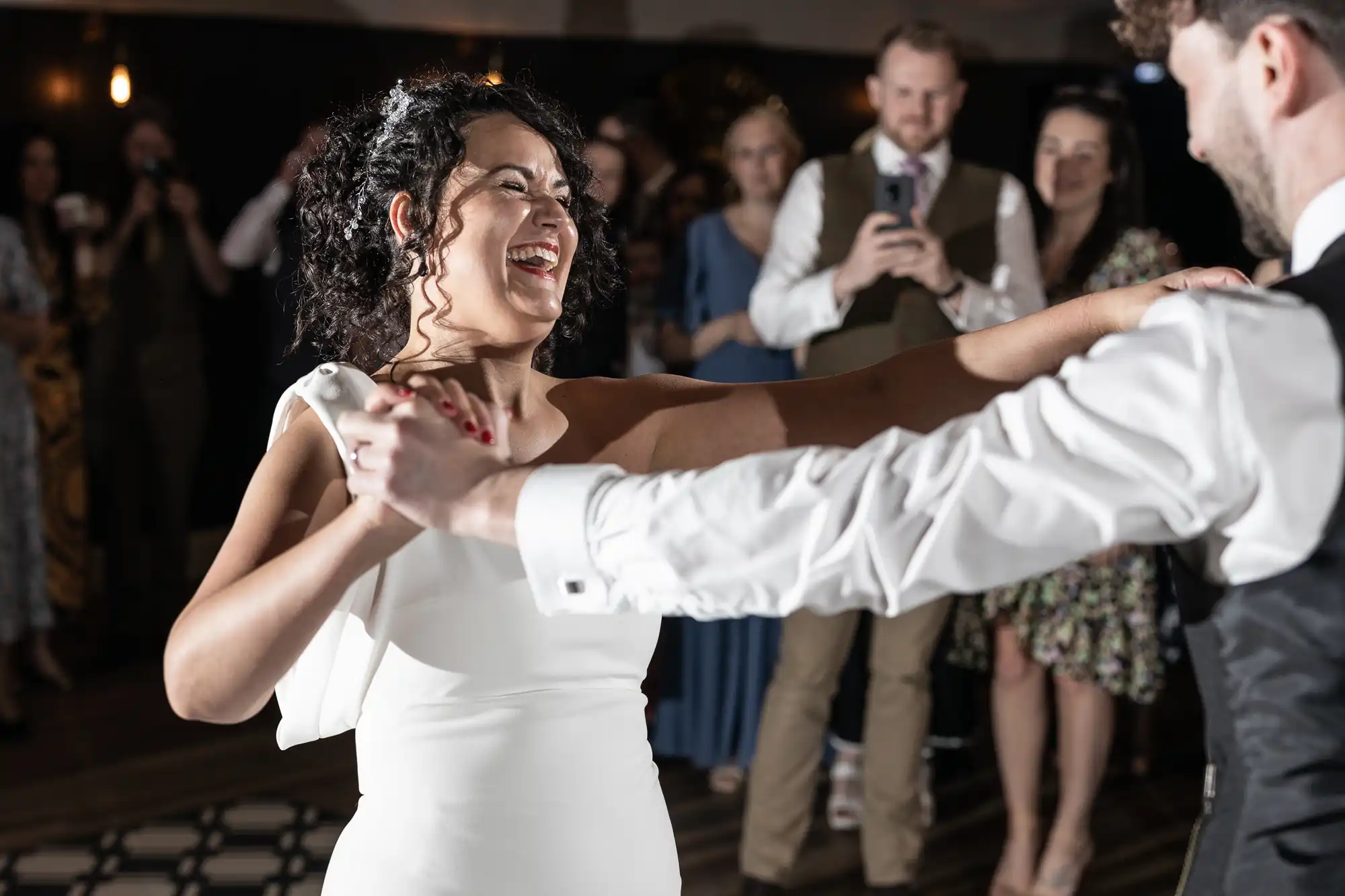 A woman in a white dress and a man in a black vest dance together, smiling, while people in the background watch and take photos.