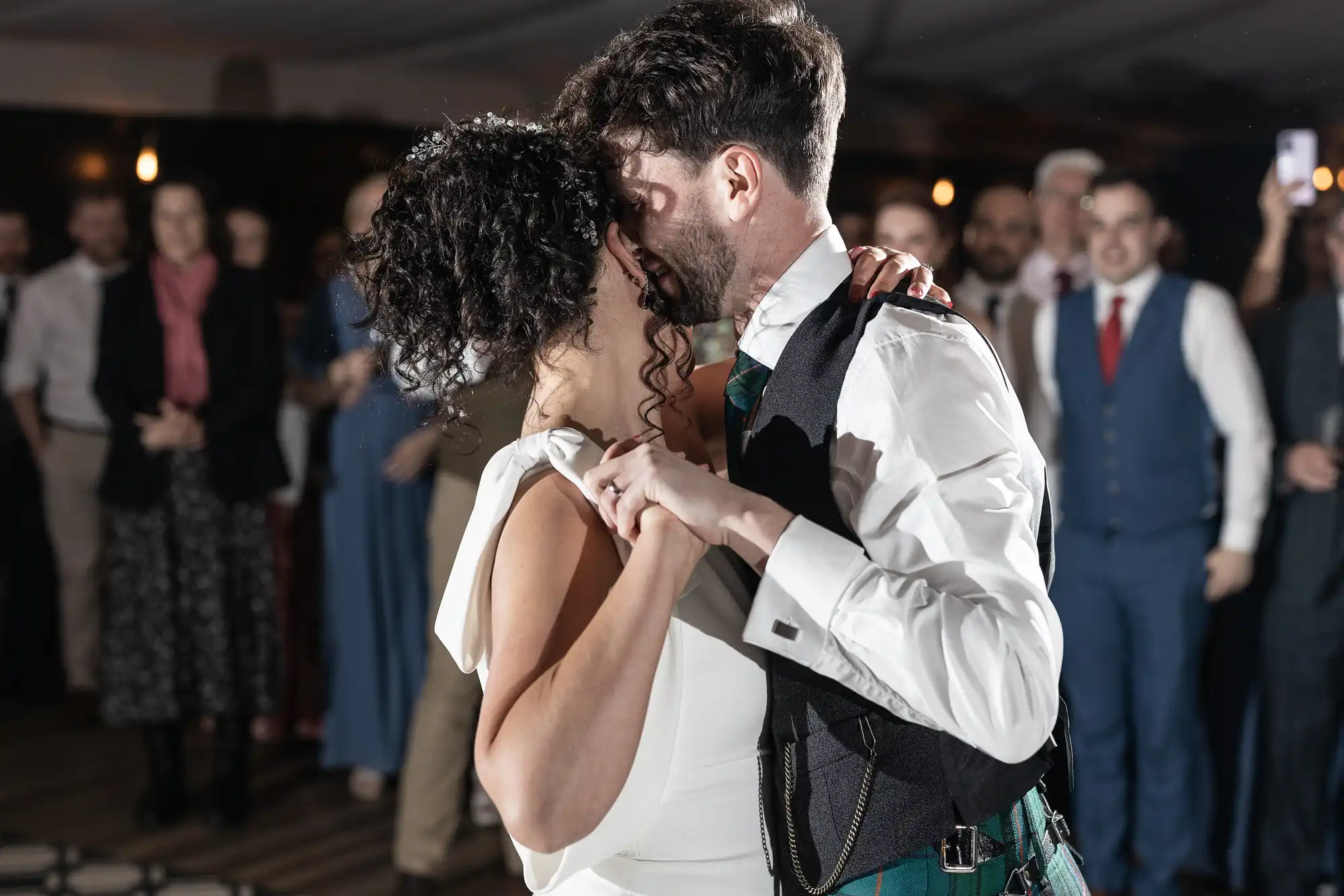 A bride and groom embrace while dancing at their wedding reception, with guests watching in the background.