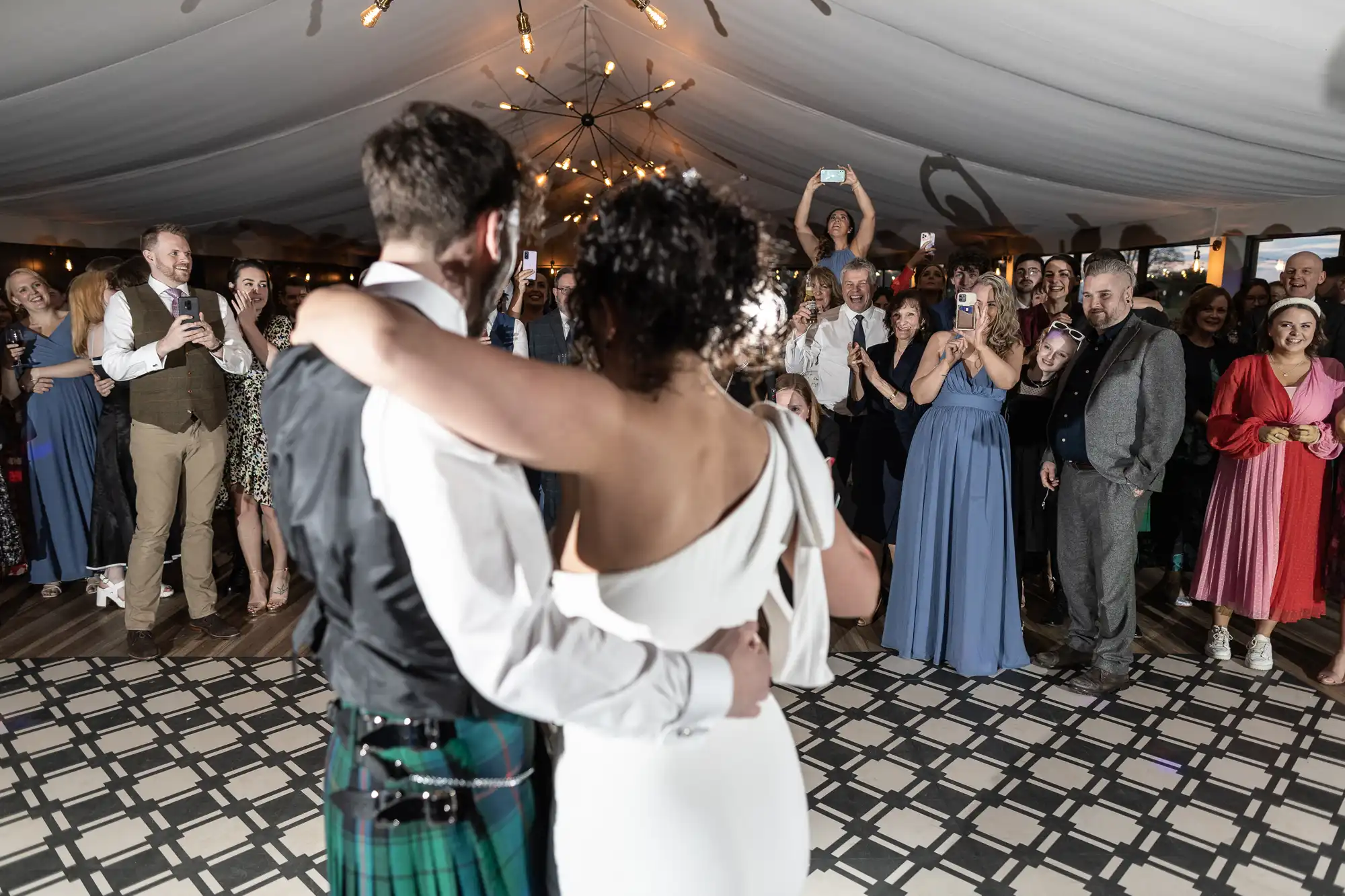 A couple embraces on the dance floor at a wedding reception, surrounded by smiling guests and well-wishers.