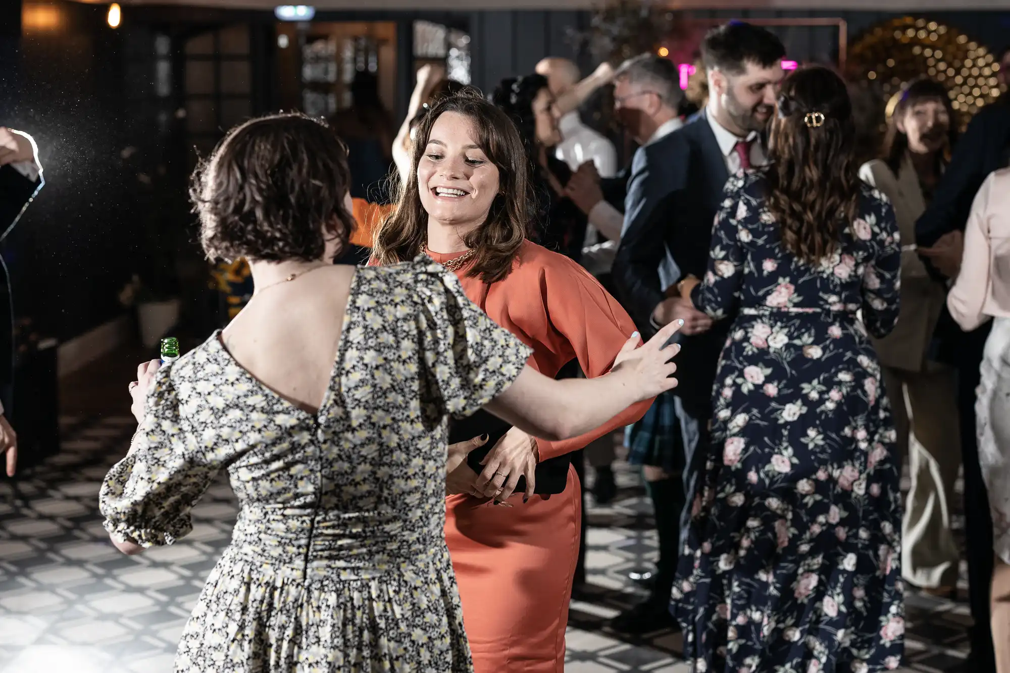 A group of people dancing in a well-lit indoor space, with two women in the foreground smiling and facing each other.