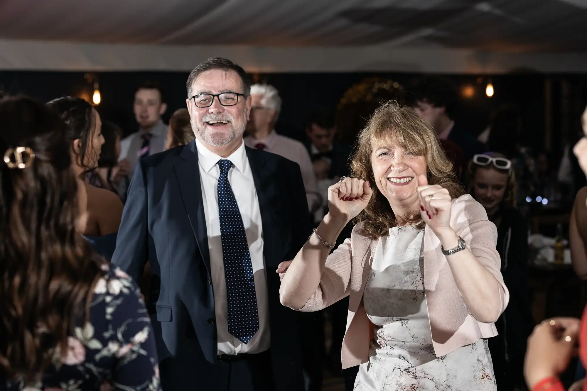 A man and a woman are smiling and dancing together at an indoor event with other people in the background.