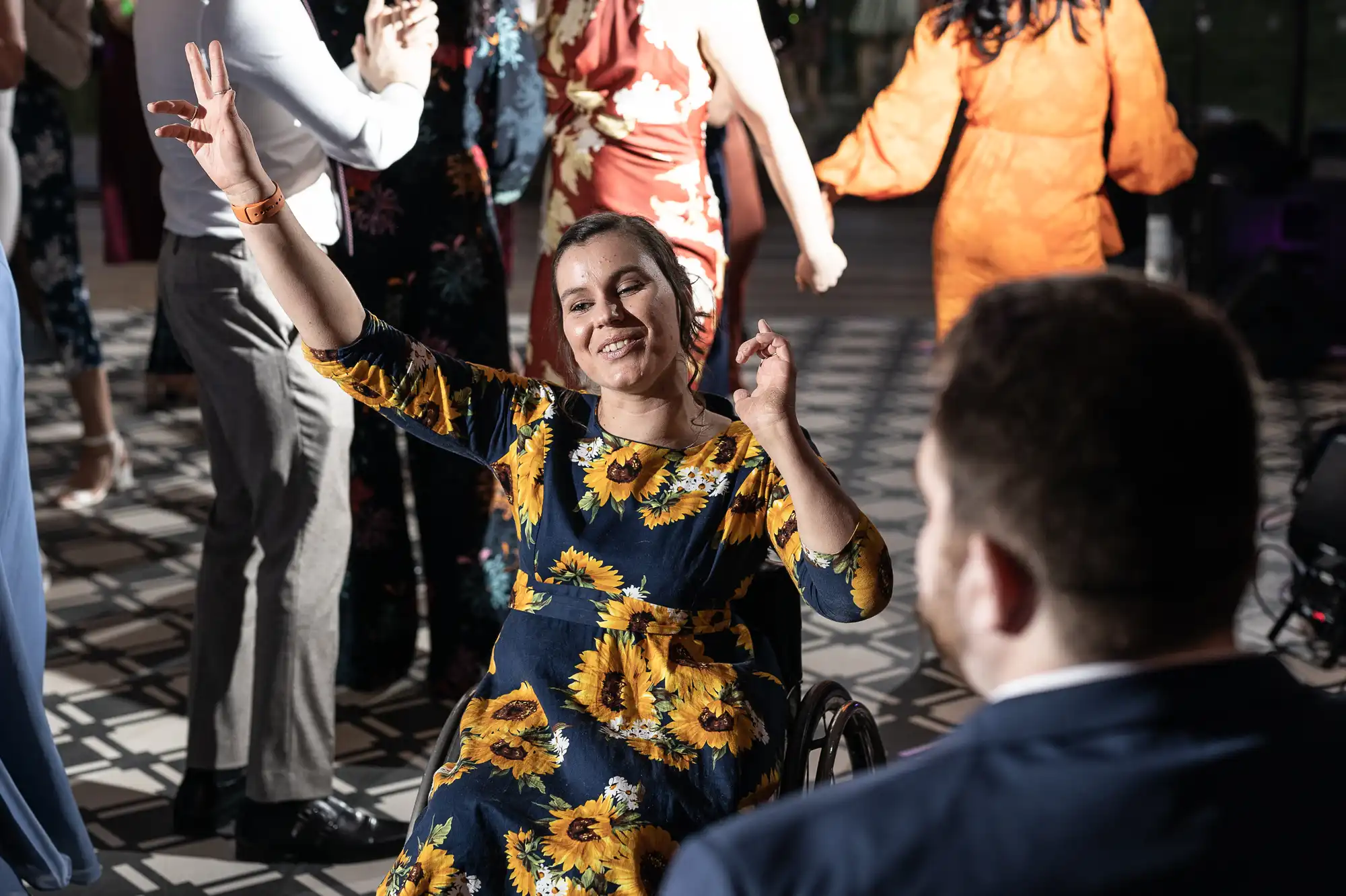 A woman in a sunflower-patterned dress and a wheelchair raises her arm while smiling at a social gathering. Other attendees can be seen dancing in the background.