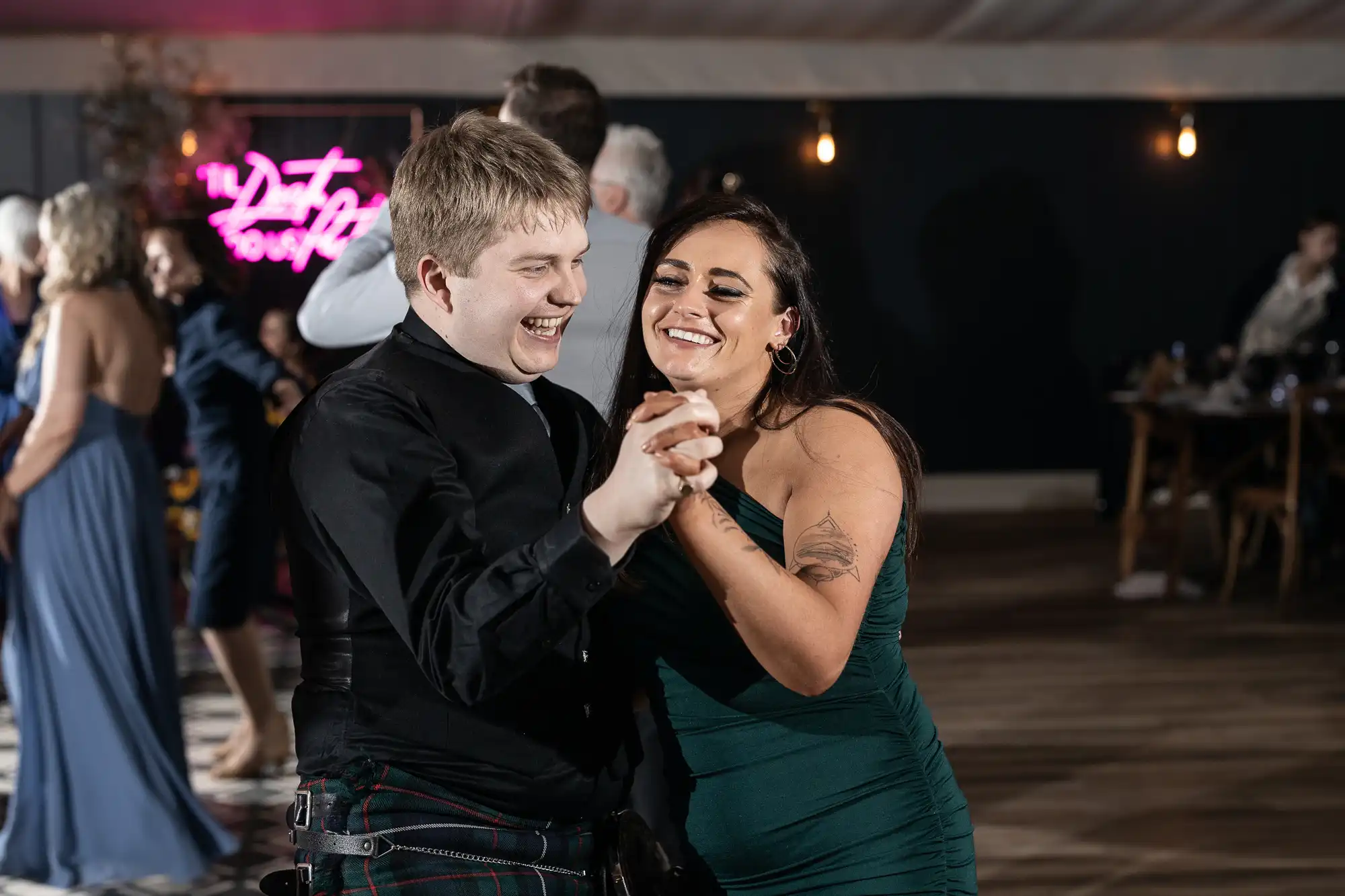 A man and a woman are dancing together at an event. The woman is in a green dress and smiling. Other guests can be seen in the background.