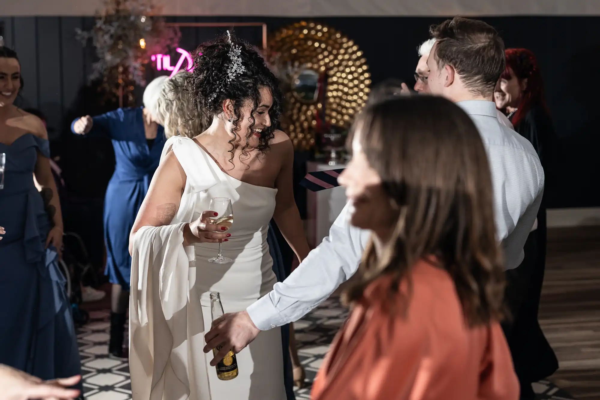 A bride in a white dress holds a glass while dancing with guests. Other individuals are in the background, one holding a beer bottle. The setting appears to be an indoor celebration or reception.