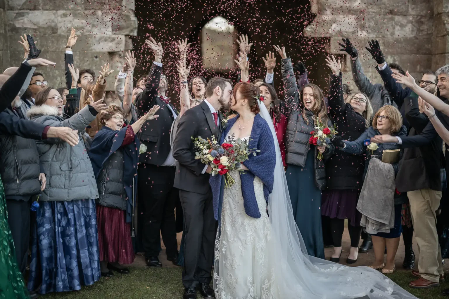 A bride and groom kiss outside as guests surround them, throwing confetti and cheering, with an old stone archway in the background.