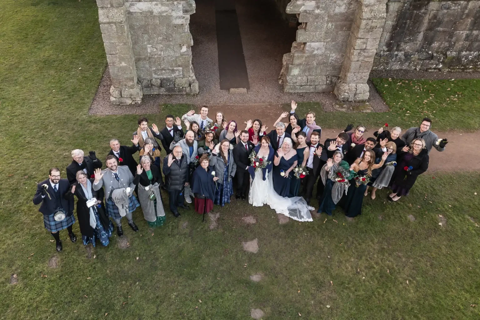 A large wedding group poses and smiles for an overhead photo outside a stone building, with the bride and groom at the center.