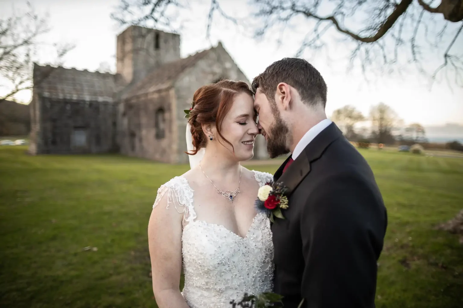 A bride and groom stand close together, touching foreheads and smiling, in front of an old stone building on a grassy lawn.