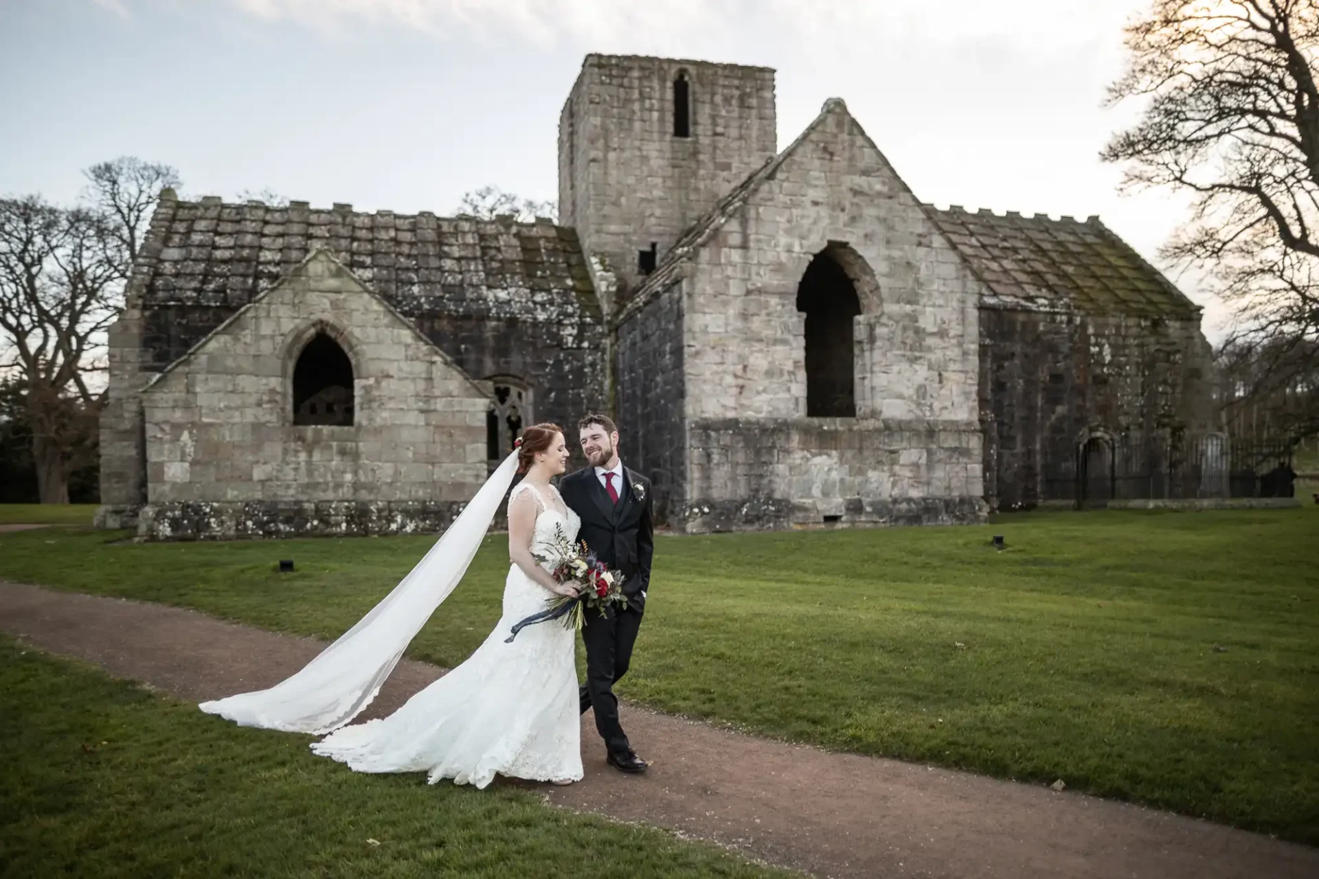 A bride and groom walk together outside an old stone church or abbey ruin, with grass and trees in the background.