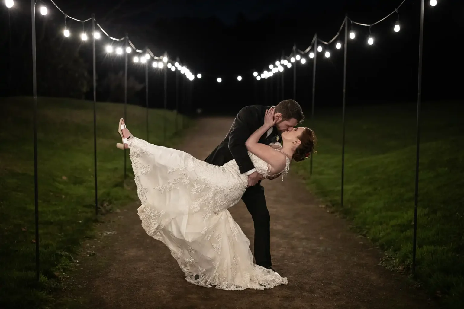 A groom dips and kisses a bride in a white gown on a path lit by string lights at night, at Dunglass Estate, East Lothian.
