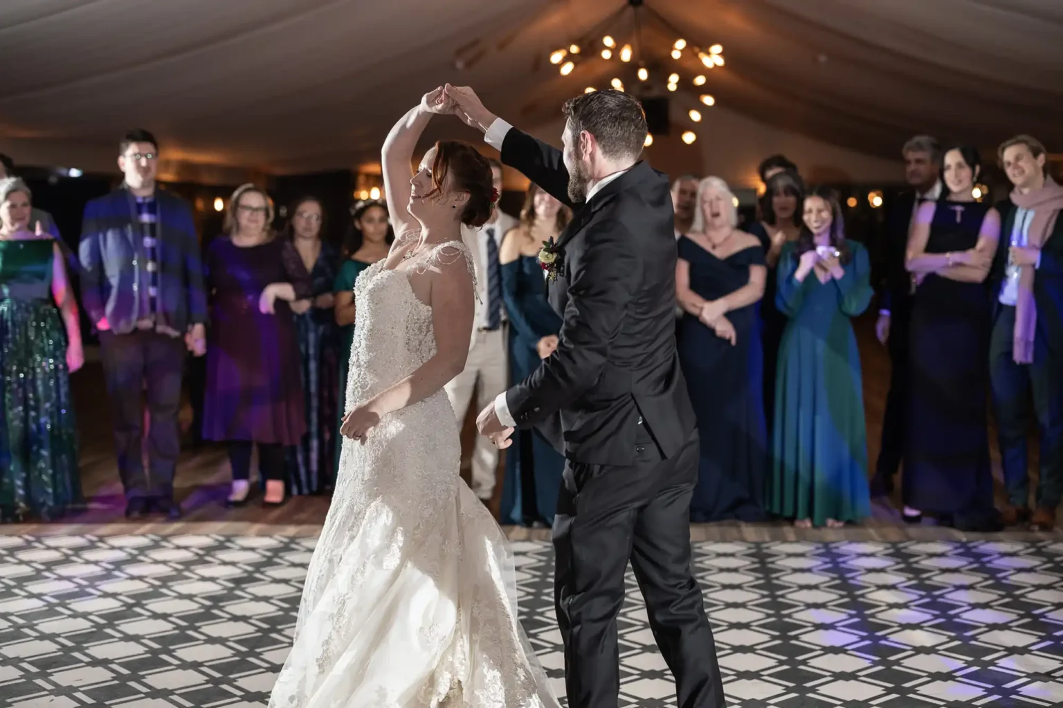 A bride and groom dance together in a formal setting, surrounded by guests who are watching and smiling.