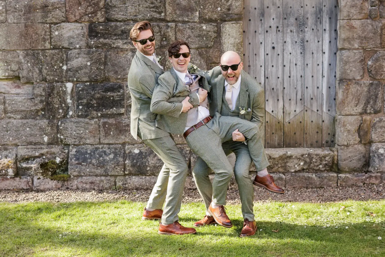 Three men in grey suits and sunglasses pose playfully in front of a stone wall and wooden door, with two lifting the third off the ground.
