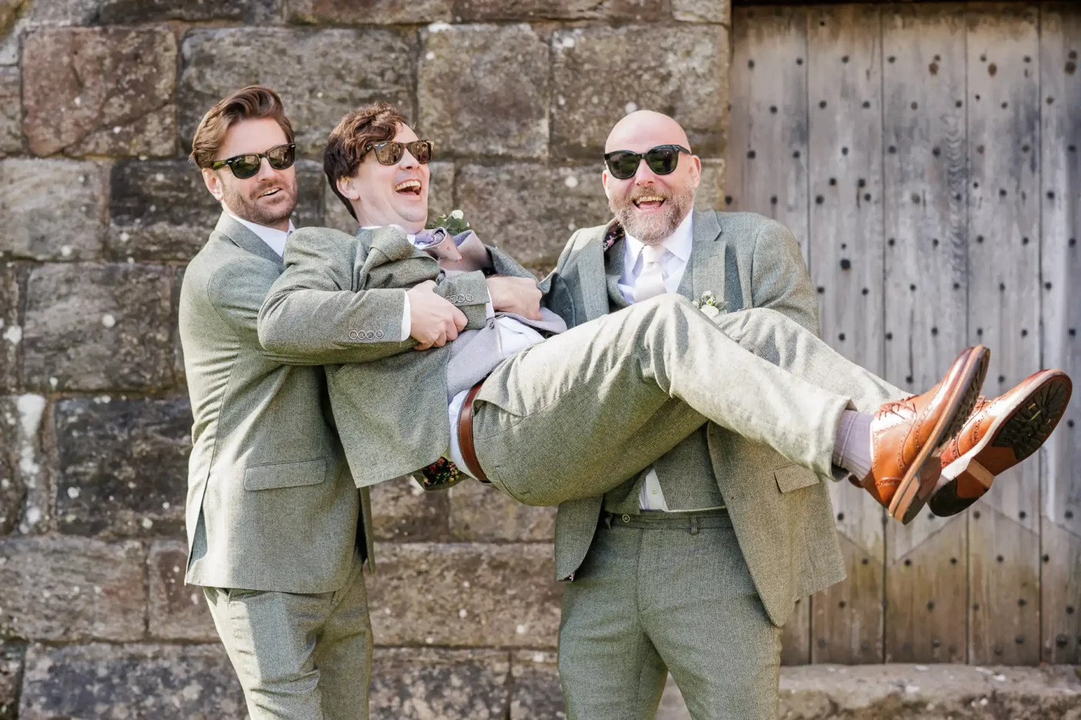 Three men in suits and sunglasses smile as two of them lift the third man in front of a stone wall and wooden door.