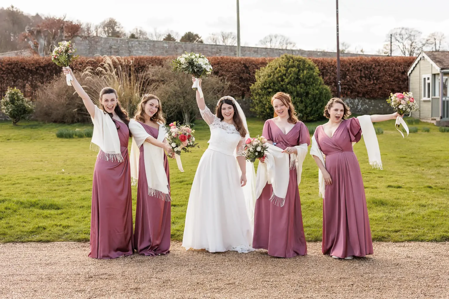 Five women, one in a white wedding dress and four in matching mauve dresses with white shawls, stand outdoors holding bouquets and smiling at the camera.