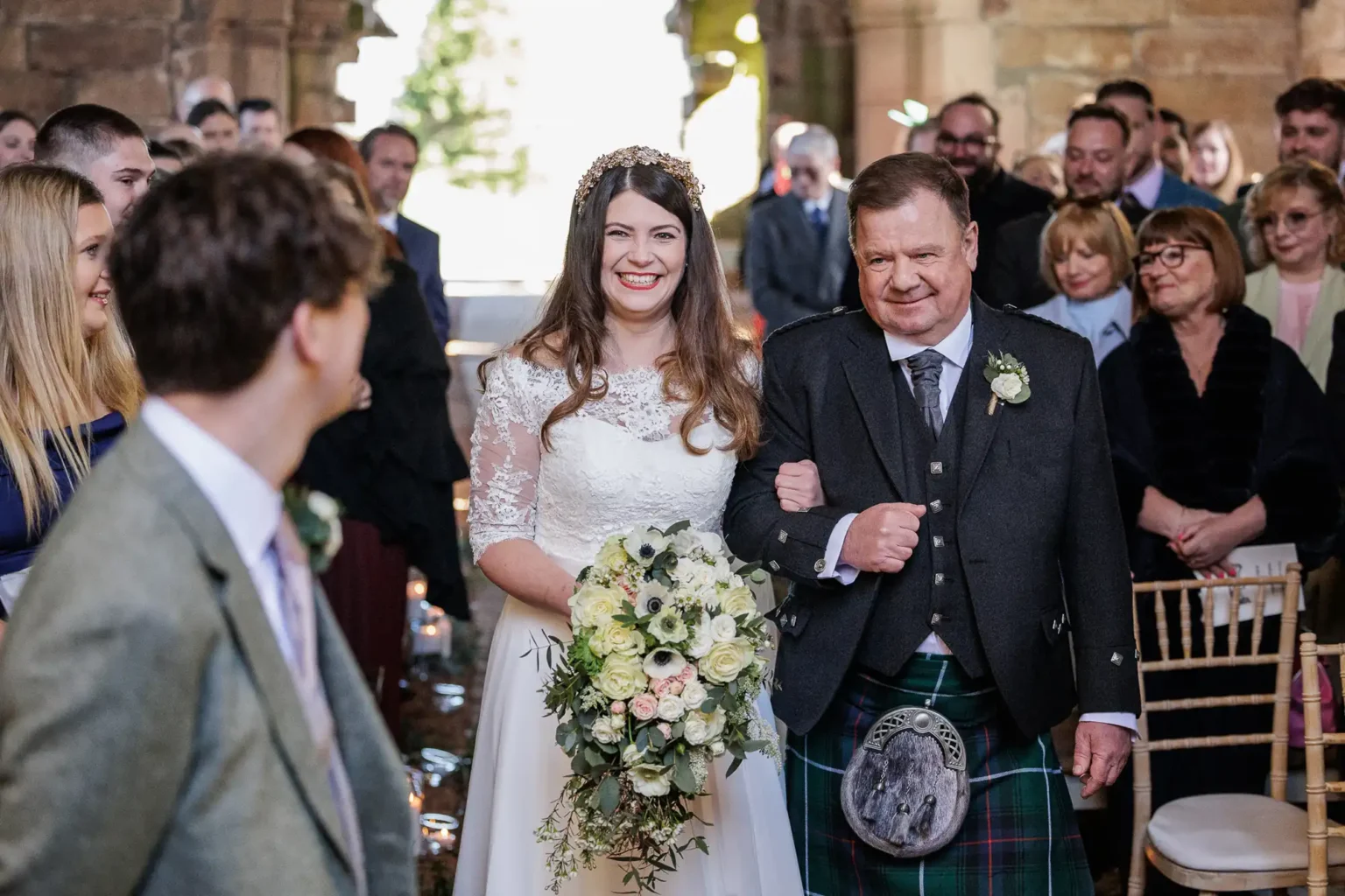 A bride in a white dress and a man in a kilt walk down the aisle, surrounded by seated and standing wedding guests in a decorated venue.