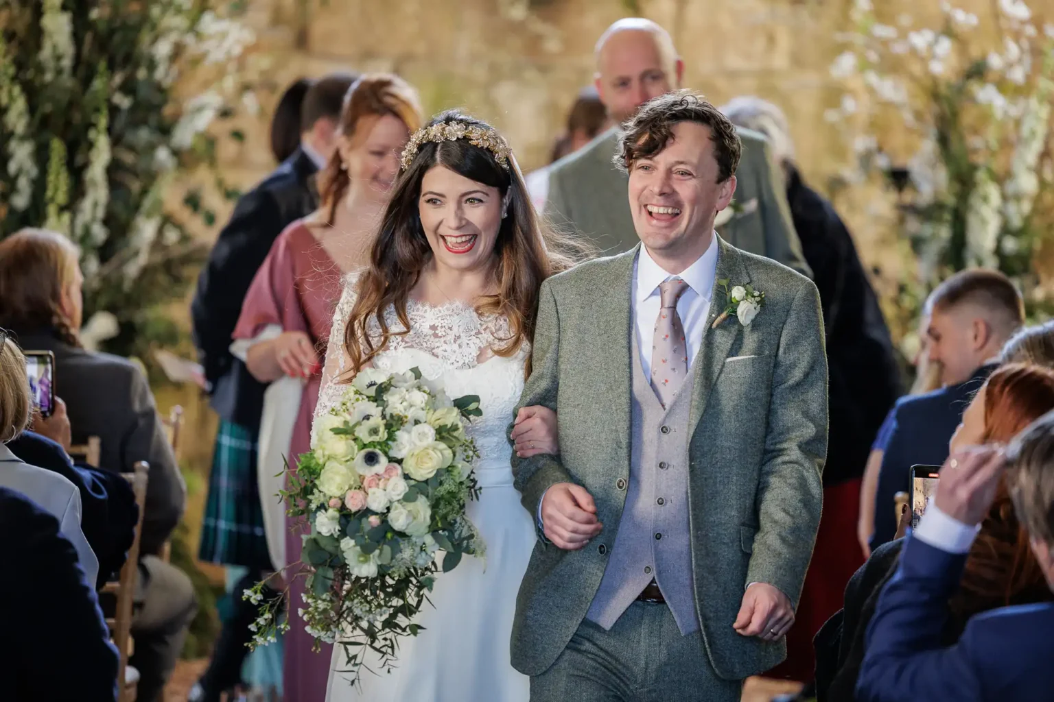 A bride and groom walk arm in arm down the aisle, smiling, surrounded by seated guests at their wedding ceremony. The bride holds a bouquet of white and pink flowers.