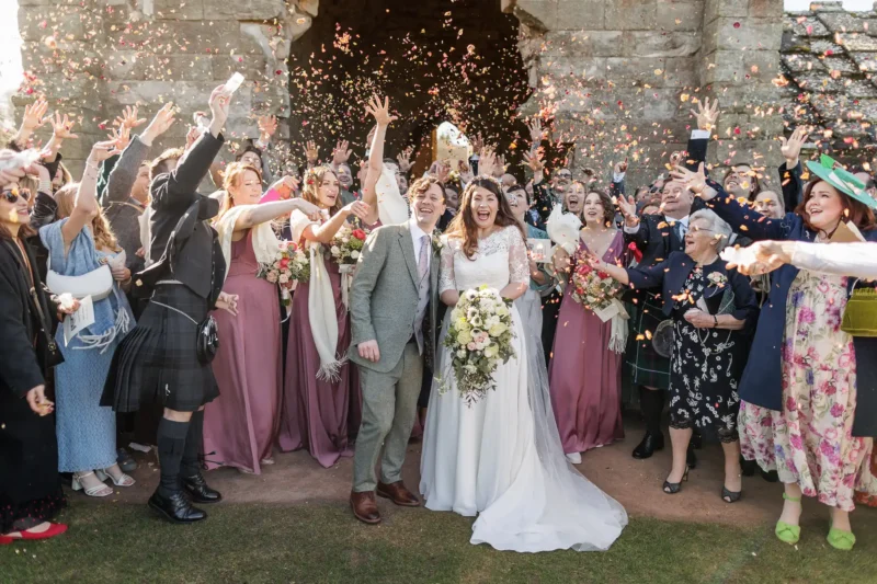 A bride and groom stand smiling surrounded by guests throwing confetti outside a stone building at a wedding celebration.