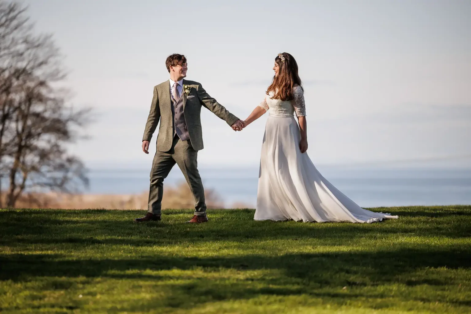 A bride and groom stand on grass, holding hands and looking at each other, with trees and water in the background at Dunglass Estate, East Lothian.