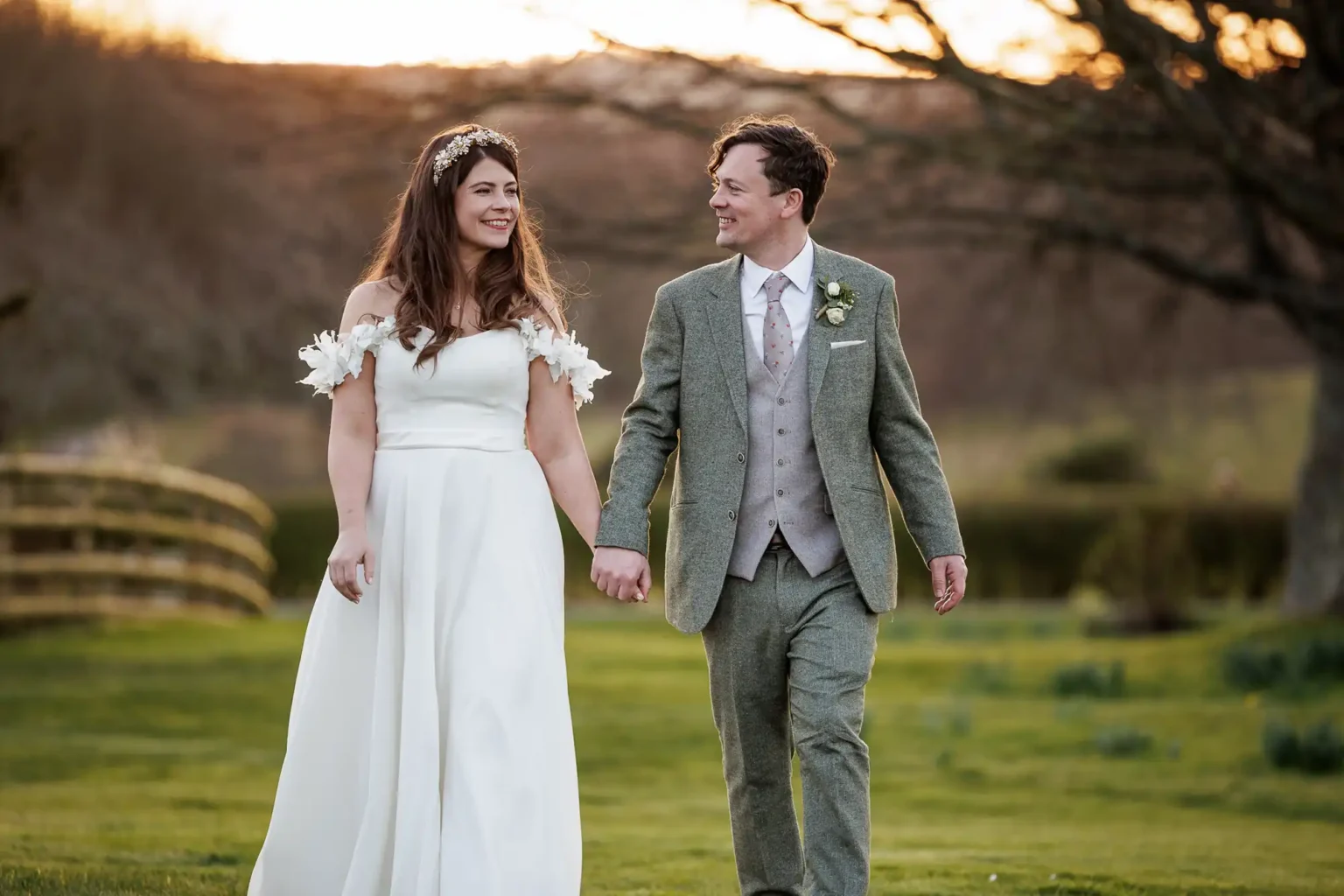 A bride in a white dress and a groom in a gray suit hold hands and walk outdoors on grass at sunset.
