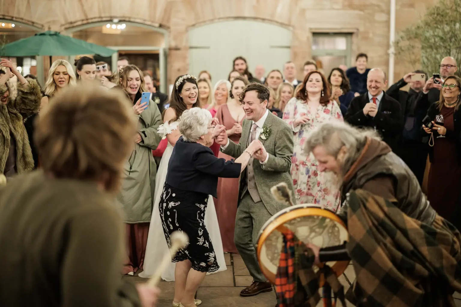 A group of people at a wedding reception watch and smile as an older woman dances with a man in a suit while a musician plays a drum.