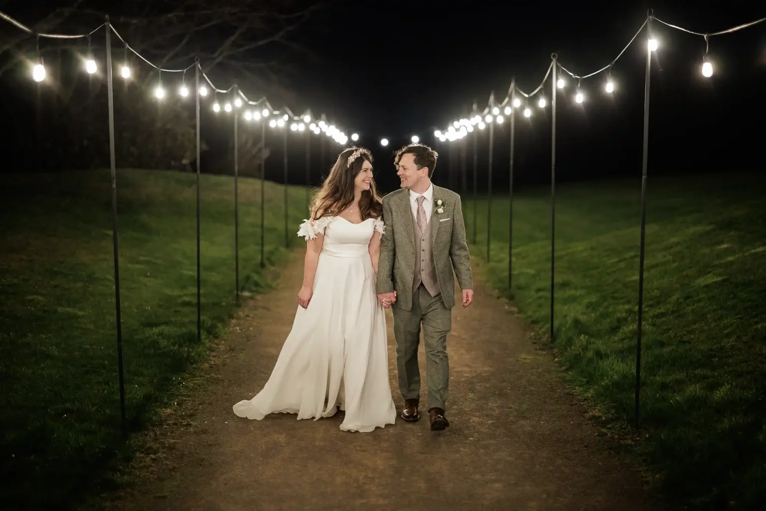 A bride and groom walk hand-in-hand down a lit pathway at night, surrounded by grass and string lights overhead.