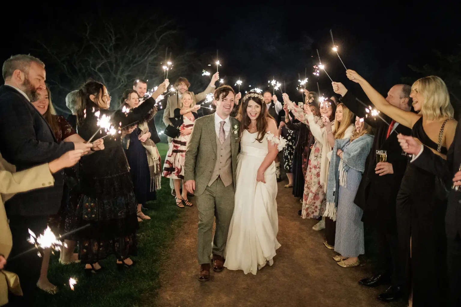 A bride and groom walk down an outdoor path at night, surrounded by guests holding sparklers and smiling as they celebrate.