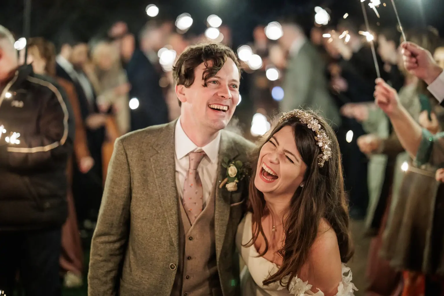 A couple in wedding attire laugh together while surrounded by guests holding sparklers at an outdoor celebration.