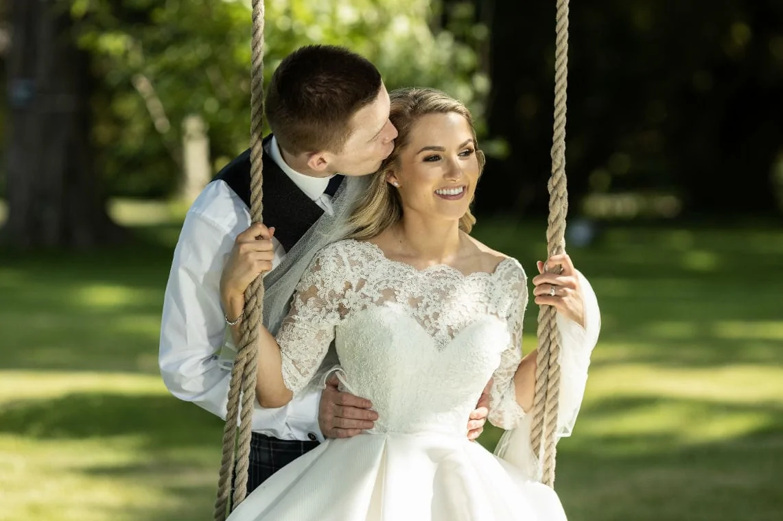 groom kisses bride as she sits on swing