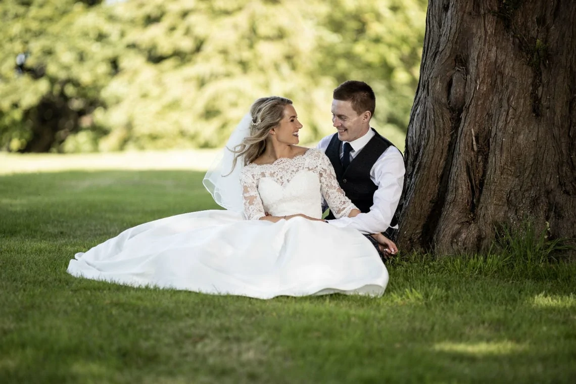 bride and groom sitting on the grass under a tree