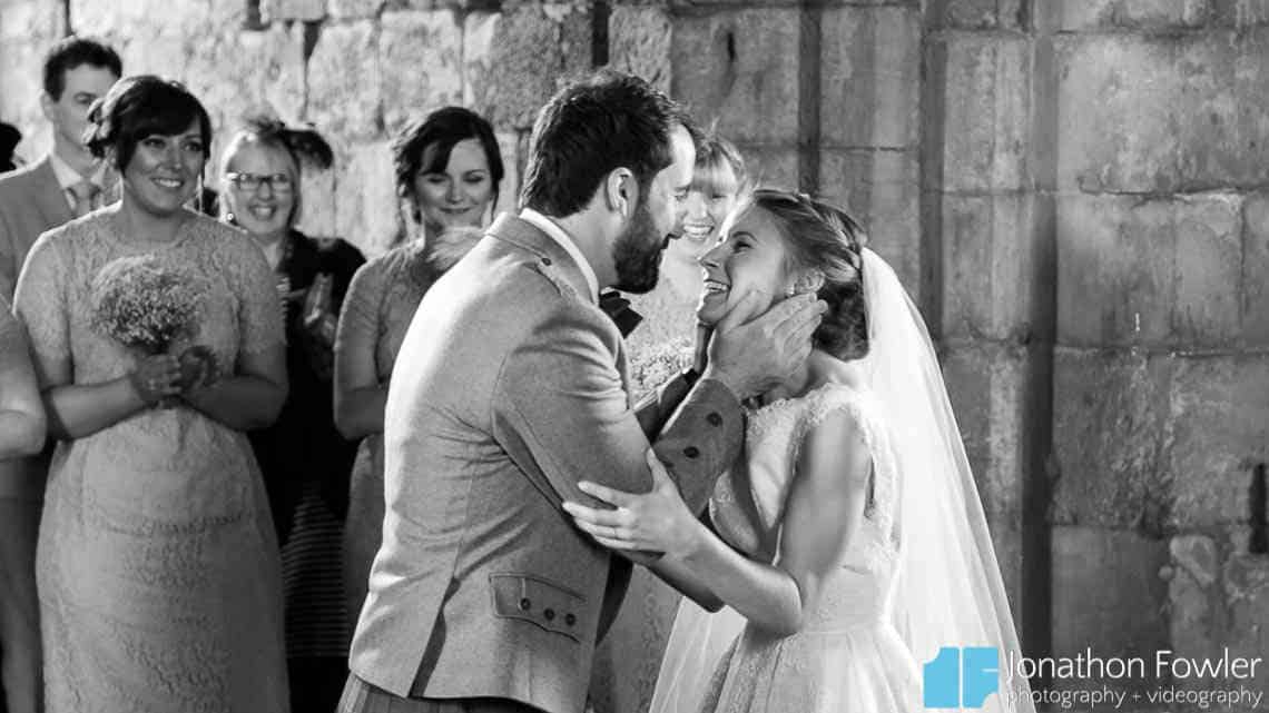 A black and white photo of a joyful bride and groom embracing and smiling at each other, with guests watching in the background.