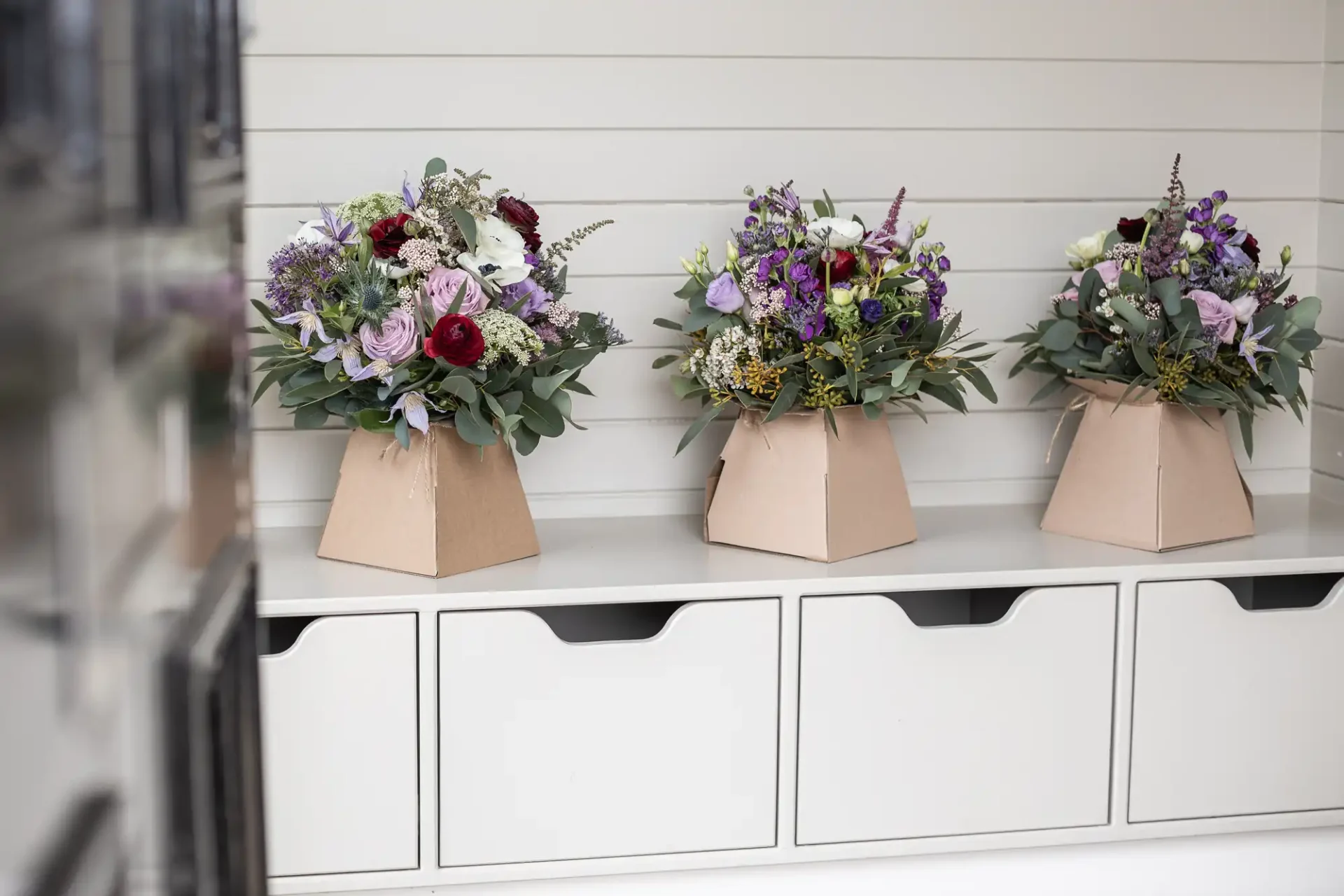 Three bouquets of mixed flowers in brown paper vases are placed on a white storage unit with drawers.