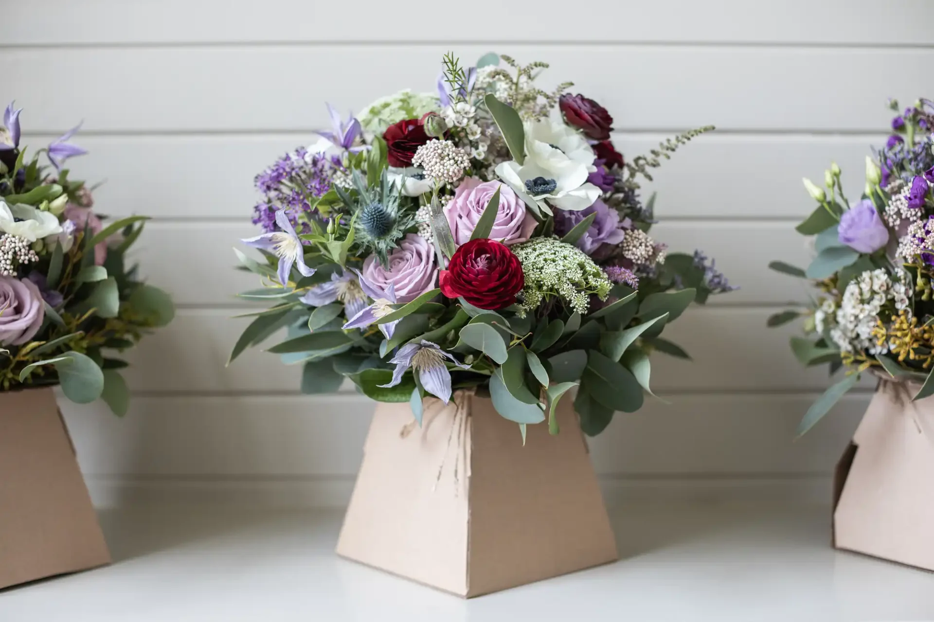 A bouquet of flowers including red, purple, and white blooms with green foliage arranged in a simple brown paper vase, set against a light-colored, horizontal wooden panel background.