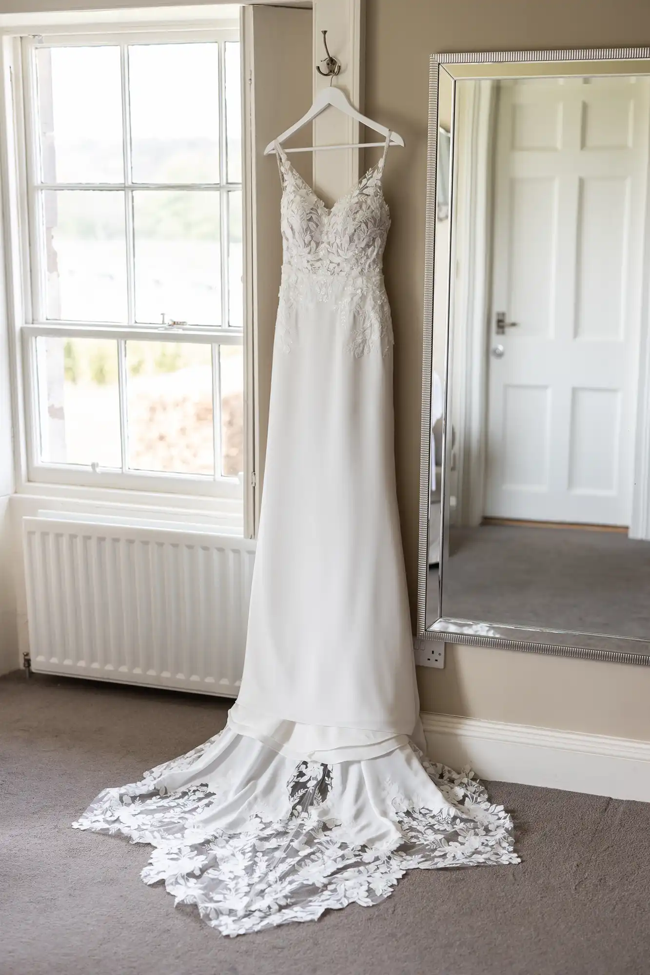 A white wedding dress with a lace bodice and a long train hangs on a hanger against a beige wall next to a large mirror.