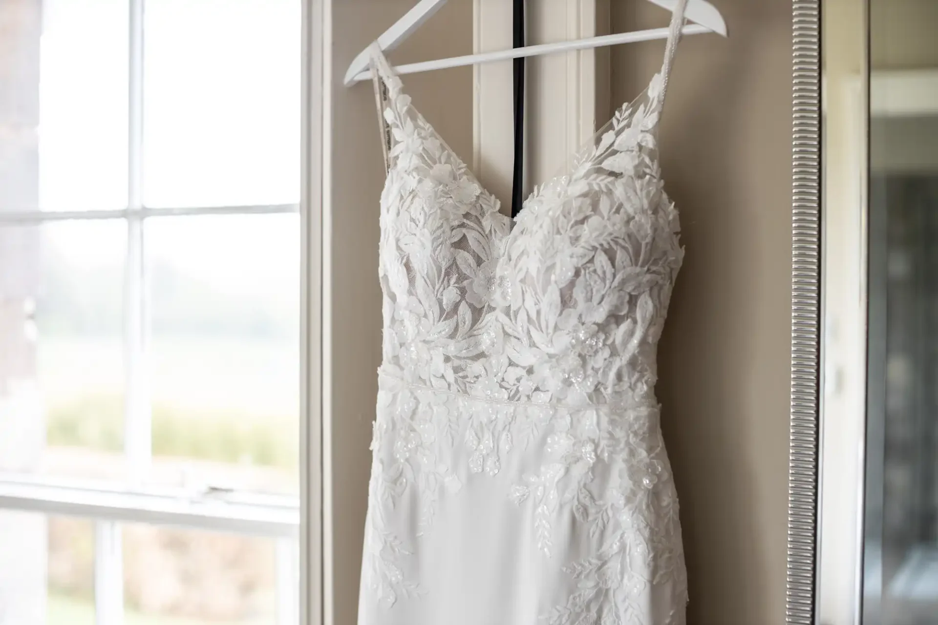 A white lace wedding dress hung on a hanger by a window with natural light streaming in.