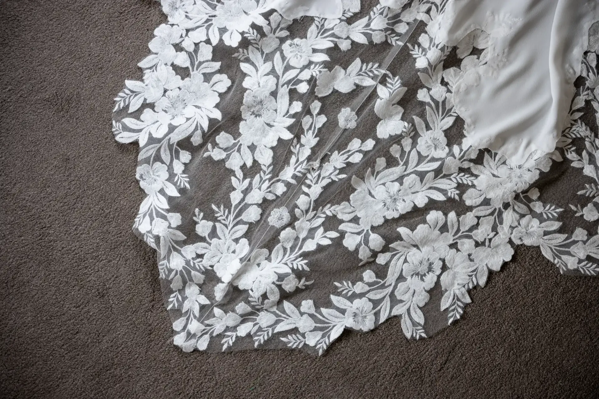 Close-up of white lace floral embroidery on a sheer fabric, laid over a grey carpet.