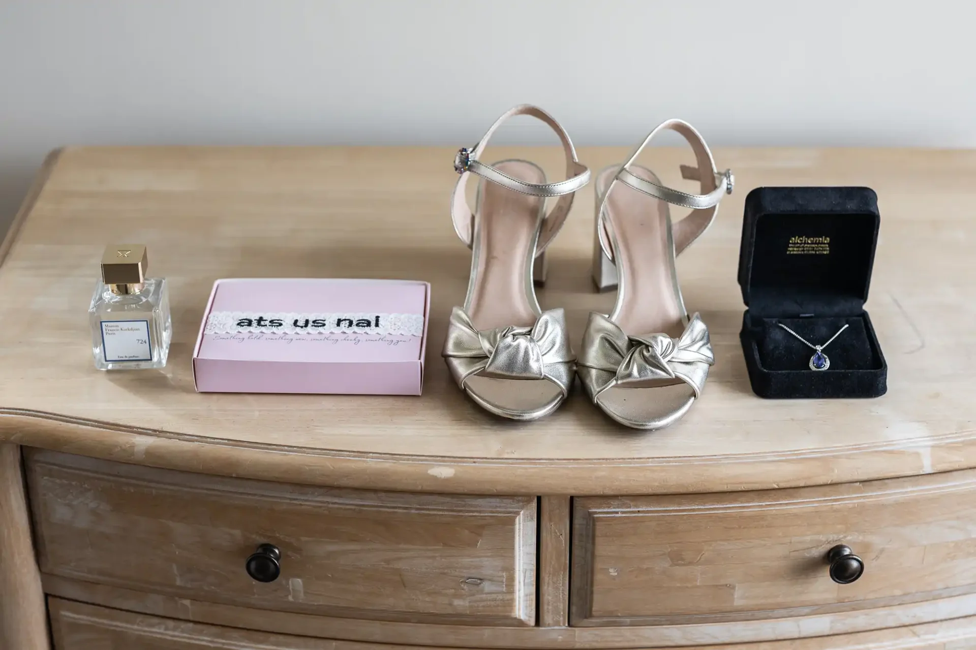 A pair of metallic high-heeled sandals placed on a wooden dresser, flanked by a perfume bottle and a small box on the left, and an open jewelry box with a necklace on the right.