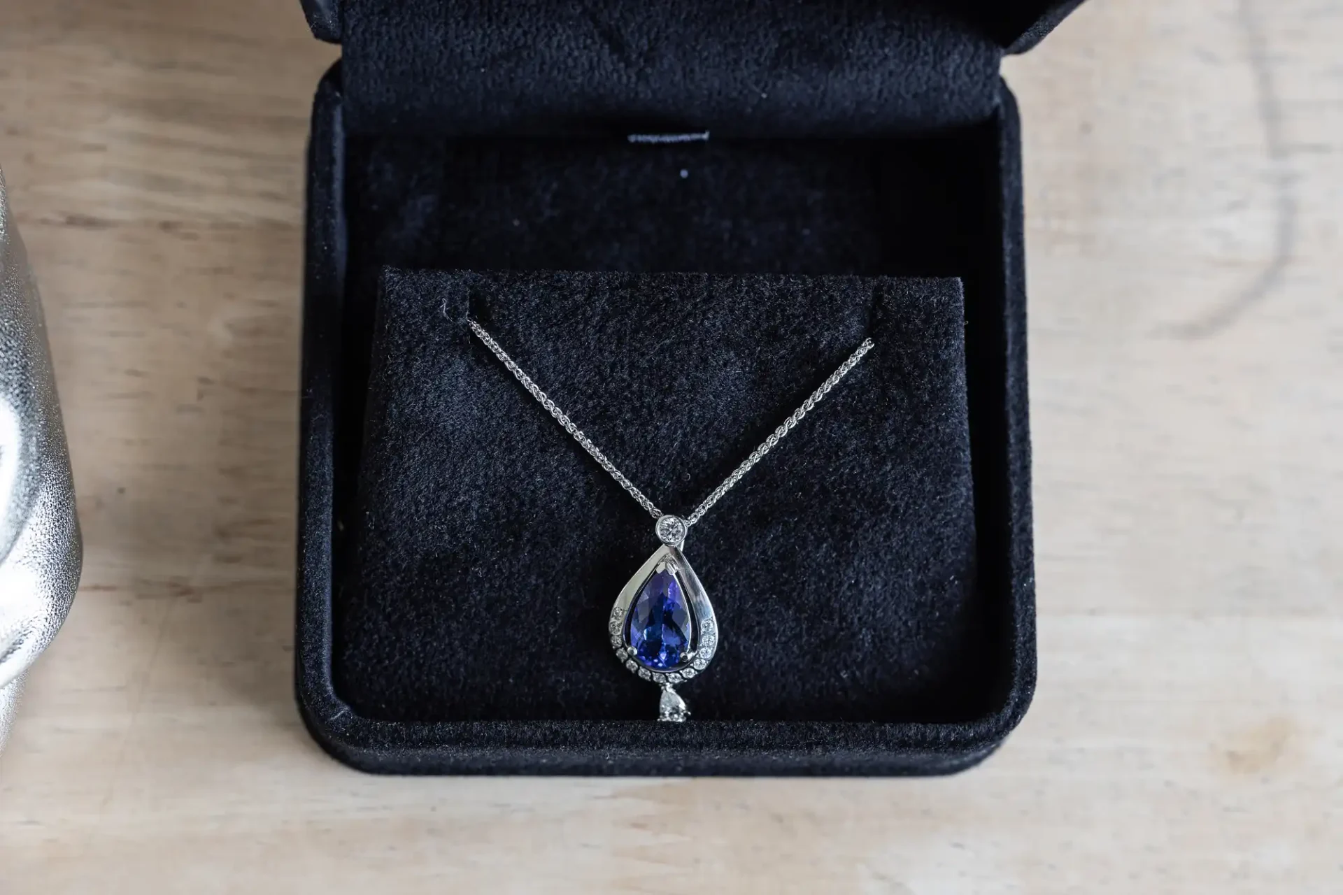 A teardrop-shaped sapphire and diamond necklace in a black jewelry box on a wooden surface.
