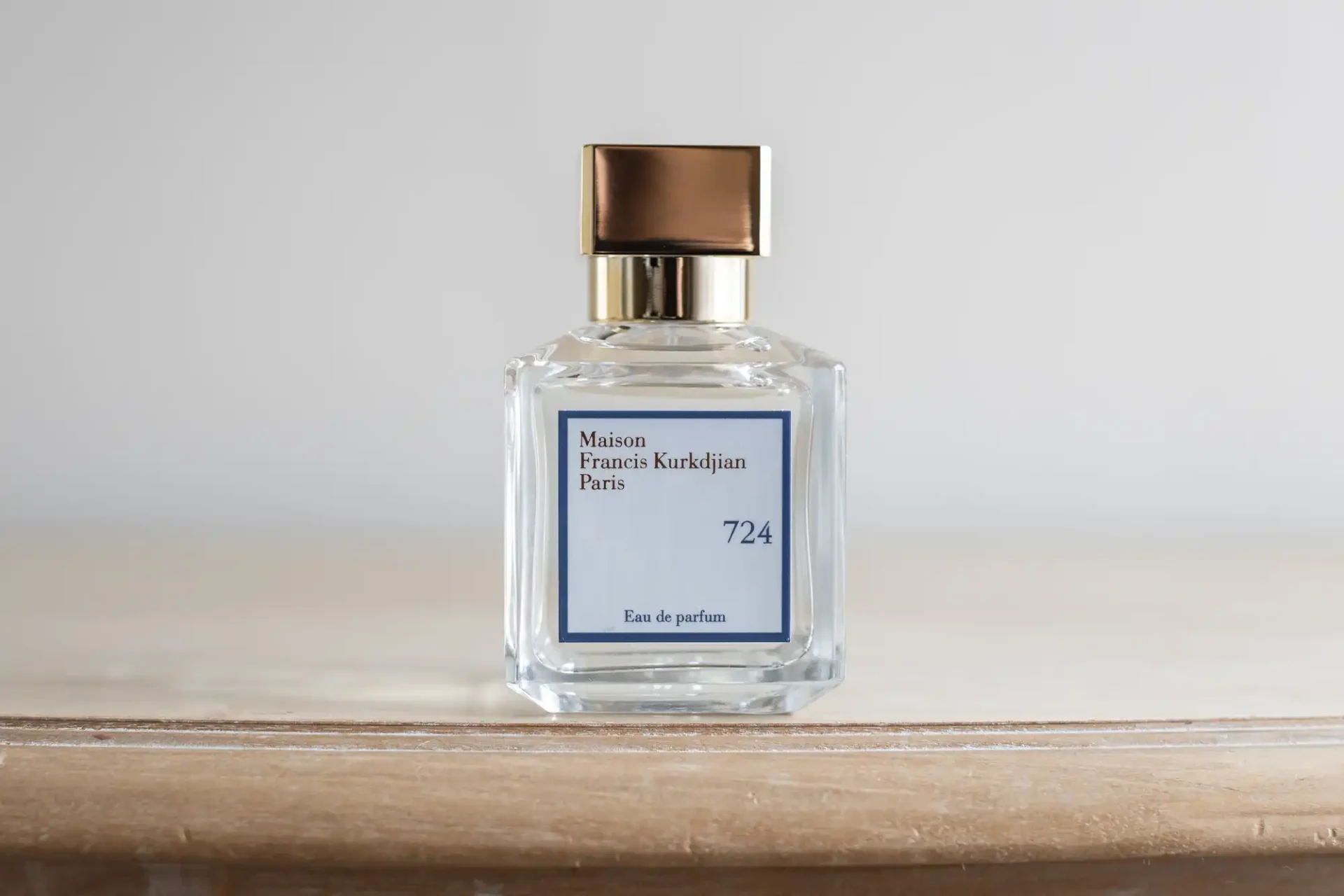 A clear glass bottle of Maison Francis Kurkdjian Paris Eau de Parfum with a gold cap, labeled "724," placed on a wooden surface.
